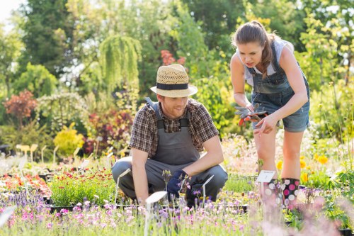 A woman and a girl are working together in a vibrant garden, planting or tending to a flower bed filled with various small, colorful flowering plants. The woman, wearing a straw hat, a checked shirt, and gardening gloves, is kneeling on the grass, closely inspecting or handling the plants. The girl, dressed in denim overalls and a light t-shirt, is standing and leaning forward, holding garden shears or a small gardening tool, actively participating in the gardening task. The garden features a lush, green environment with mature trees and shrubs in the background, creating a serene outdoor space typical of South Kensington. The flower bed is bordered by a low, natural looking edge, and the surrounding lawn area appears well-maintained with dense, healthy grass. Soft sunlight filters through the trees, casting gentle shadows and enhancing the natural tones of the plants and soil. This scene exemplifies gardening activities supported by professional landscaping services such as those offered by Landscapers South Kensington, emphasizing outdoor leisure and horticultural care in a well-kept residential garden in the area.