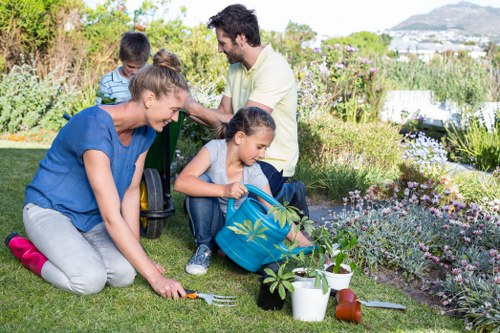 A family of four engaging in gardening activities in a well-maintained backyard garden, with a young girl planting a small shrub in the grass while a woman with a gardening fork assisting nearby. In the background, a man and a boy observe the scene, surrounded by lush green plants, flowering shrubs, and potted plants placed along the edge of the flower bed. The lawn is lush and evenly mown, bordered by a variety of flowering perennials and evergreen shrubs, contributing to the vibrant and neat outdoor space. The garden features a mix of paved pathways and grassy areas, with sunlight illuminating the scene under clear weather, highlighting the natural tones of the garden's flora and horticultural elements. This setting reflects a well-kept outdoor environment suitable for landscaping and garden care services provided by South Kensington-based landscapers, emphasizing active involvement in gardening, planting, and outdoor maintenance, with a backdrop suggesting proximity to natural and urban landscape features common in South Kensington or nearby areas.
