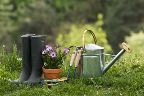 The image displays a neatly maintained garden area with a lush green lawn France, featuring seasonal flowering plants in a small terracotta pot, with vibrant purple and white blossoms. Nearby are a pair of black rubber gardening boots placed on the grass, indicating active outdoor work. The background includes a softly blurred mix of trees and shrubs, suggesting a natural, well-established garden environment typical of South Kensington. In the foreground, garden tools such as a garden fork and pruning shears rest against a metal watering can with a curved spout, highlighting ongoing gardening activities. The scene is lit by natural daylight with clear weather, emphasizing the healthy greenery and flower colours. The garden layout is simple yet functional, with the tool and plant arrangement making it suitable for home gardening or landscaping services offered by Landscapers South Kensington in the local area. This outdoor space reflects a typical suburban garden that benefits from professional gardening and outdoor maintenance services, creating a welcoming environment for relaxed outdoor activities.