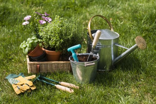 A neatly maintained outdoor garden area in South Kensington featuring a lush, green lawn with evenly trimmed grass. In the foreground, there is a collection of gardening tools and supplies, including a pair of yellow gardening gloves, small hand trowels with wooden handles, and a pair of pruners. To the left, a rectangular wicker basket holds potted plants with green foliage and purple flowers, adding a splash of natural colour. Behind these, there is a silver metal watering can with a wooden handle positioned on the grass, alongside a smaller silver bucket containing soil and gardening tools. The background reveals a landscape with evenly spaced hedges or shrubs bordering the garden, creating a structured outdoor space suitable for landscaping and garden maintenance services. The scene is well-lit under natural sunlight, suggesting a clear day, and the overall environment reflects a tidy, cultivated garden typical of residential properties serviced by professional landscapers in areas like South Kensington or nearby districts, emphasizing the company's expertise in outdoor garden care and landscaping in the London region.