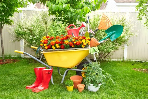 A vibrant backyard garden with a well-maintained lawn bordered by a white fence and lush green trees providing shade. Central to the image is a yellow wheelbarrow filled with blooming red and orange flowers, positioned on a neatly cut grassy area. Surrounding the wheelbarrow are gardening tools and accessories, including a red watering can hanging from the handle, a small potted plant placed beside it, and a pair of bright red rubber boots on the grass. To the right, a white pot with leafy green plants sits alongside several empty terracotta plant pots of different sizes, ready for planting. In the background, dense foliage and tall shrubs contribute to a lush outdoor environment, with the sunlight filtering through the leaves, indicating a clear and pleasant day. This scene reflects typical outdoor maintenance activities, suitable for gardening and landscaping services in South Kensington, with a focus on outdoor aesthetic enhancement and plant care.