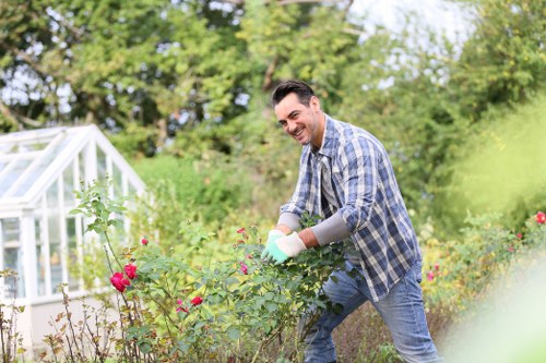 A man with dark hair, dressed in a plaid shirt and jeans, is working in a garden surrounded by lush greenery and flowering bushes, including pink roses in the foreground. He is smiling while tending to the plants, wearing gardening gloves, and appears to be pruning or inspecting the shrubs. The garden features a well-maintained lawn area, bordered by flower beds and natural soil patches. In the background, there are mature trees with dense green foliage, providing shade and a natural backdrop. Part of a white greenhouse structure with glass panels is visible on the left side of the image, indicating an outdoor space dedicated to gardening and plant care. The scene is set during daylight hours with bright, natural lighting, reflecting a peaceful, well-kept outdoor environment typical of private garden spaces in South Kensington, Surrey. This image reflects professional gardening and lawn maintenance activities that a company like Landscapers South Kensington might provide, highlighting elements such as healthy plants, clean gardening tools, and a serene outdoor setting.