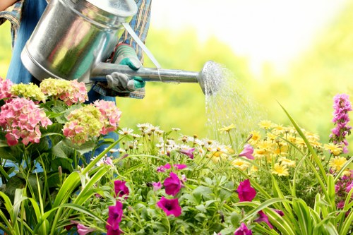 A person wearing gardening gloves is watering a flower bed in an outdoor garden area. The bed contains a variety of plants, including pink and yellow flowers, with green foliage and tall grass-like plants at the forefront. The background shows a lush, green landscape with trees and natural light, suggesting a bright and pleasant day. The scene reflects typical gardening activities that a landscaping company like Landscapers South Kensington might undertake to maintain and beautify outdoor spaces in London or surrounding areas. The garden layout features a mix of flowering plants and dense greenery, with natural soil and paved pathways partially visible, indicating a well-designed landscaped yard suitable for outdoor household gardens or private gardens in South Kensington.
