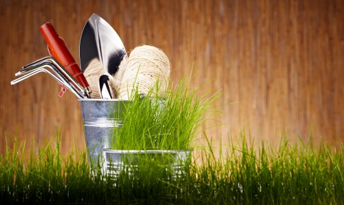 The image features a close-up view of gardening tools and materials placed on a grassy surface with a blurred wooden fence background. In the foreground, vibrant green grass covers the ground, providing a natural setting. Behind the grass, a small metal bucket holds various gardening tools, including a trowel and a hand rake, with wooden handles visible. Next to it, there is a ball of natural twine or garden string for tying plants or securing garden structures. The setting suggests a prepared outdoor space, likely a garden area ready for planting or maintenance, with natural lighting indicating a clear day. This scene highlights essential gardening equipment and the lush, well-maintained appearance of a typical outdoor garden environment, supporting professionals such as those from Landscapers South Kensington in providing comprehensive gardening and landscaping services in the local area, including South Kensington and surrounding regions.