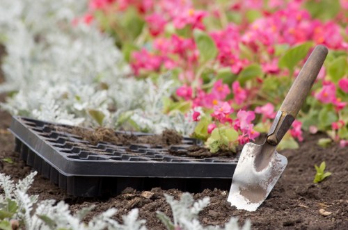 A gardening scene in a residential outdoor garden, featuring a section of freshly turned soil bordered by a paved pathway. In the foreground, there is a black seedling tray with small soil-filled compartments, partially covered with soil and plant debris. A small hand trowel with a wooden handle and metal blade is inserted into the soil, indicating recent planting or garden preparation activities. Surrounding the area, vibrant pink and white flowering plants provide contrast to the dark, rich soil. The background hints at a well-maintained landscaped yard with a mix of flowering shrubs and greenery, under natural daylight with soft, diffuse lighting. This scene exemplifies professional gardening and planting work suitable for a residential garden in South Kensington, aligning with services related to garden maintenance, planting, and outdoor space enhancement by Landscapers South Kensington.