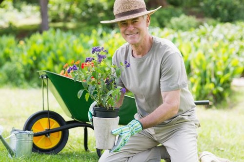 The image shows an elderly man kneeling on a well-maintained grassy lawn in a garden, holding a potted flowering plant with purple, red, and yellow blooms. He is wearing a beige hat, light grey t-shirt, and gardening gloves, smiling at the camera. Behind him is a green garden wheelbarrow with a yellow wheel, filled with gardening tools and supplies, and nearby are garden beds edged with soil and some small shrubs. The background features lush green trees and hedges, indicating a spacious outdoor area with natural light and pleasant weather, suitable for garden maintenance and landscaping work. This scene exemplifies outdoor gardening and lawn care activities that a professional landscaper such as Landscapers South Kensington may undertake in areas like South Kensington or nearby in London, focusing on garden beautification and upkeep.