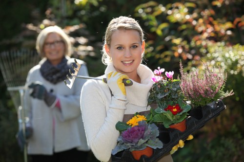 A young woman in a white sweater and gardening gloves holds a black tray filled with colorful flowering plants, including pink, purple, yellow, and red blooms, standing in a well-maintained garden during daylight with natural sunlight. Behind her, an older woman dressed similarly with a gardening tool in hand, slightly out of focus, suggests a garden gardening activity. The garden features lush green trees and shrubbery, with a mix of vibrant foliage and natural earthy tones, including a neatly trimmed lawn and flower beds bordered by soil. The background shows sunlight filtering through trees, creating a warm and inviting outdoor environment consistent with professional garden maintenance services in South Kensington. The scene reflects a tidy and cultivated outdoor space, indicative of landscaping and gardening work often provided by Landscapers South Kensington, integrating natural beauty with well-maintained plant displays.