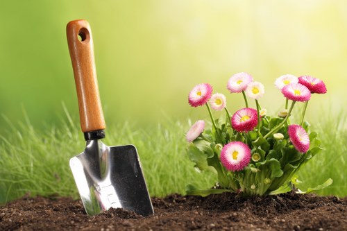 A landscaped garden scene featuring a close-up view of a section of soil with a small garden trowel, having a wooden handle and metal blade, partially inserted into the ground. To the right, a cluster of pink and white flowering plants with rounded blooms and green foliage is visible. The background displays a blurred, lush green lawn area, suggesting a well-maintained outdoor space typical of a residential garden in South Kensington. The garden appears to be in the process of planting or soil preparation, emphasizing gardening activities related to outdoor maintenance and landscape enhancement. Lighting indicates a bright, natural outdoor environment, consistent with a mild weather day. This scene aligns with gardening services offered by landscapers in South Kensington, focusing on lawn care, planting, and garden structuring.