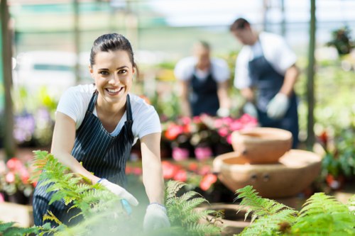 A young woman with dark hair tied back, wearing a white t-shirt and a navy blue striped apron, is working in a greenhouse or garden centre environment, tending to lush green ferns and other foliage plants. She is smiling and facing the camera while carefully handling the plants in a bright, well-lit space with large glass panels overhead, allowing natural sunlight to illuminate the area. Behind her, two other gardeners, a man and a woman, are slightly out of focus; the man is wearing an apron and is engaged in arranging or inspecting potted plants, while the woman is also involved in plant care tasks. The setting includes a variety of colorful flowering plants and structured plant displays, with some decorative ceramic fountains or water features visible in the background. The overall scene depicts professional gardening or landscaping staff maintaining healthy, vibrant garden plants, highlighting expertise in horticulture and outdoor plant care, relevant to services offered by Landscapers South Kensington in the London area.